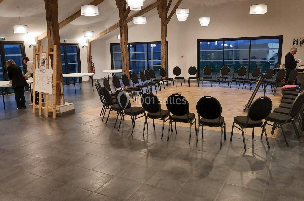 Salle avec des chaises disposées en cercle autour d'une estrade en bois, tables et personnes en arrière-plan.