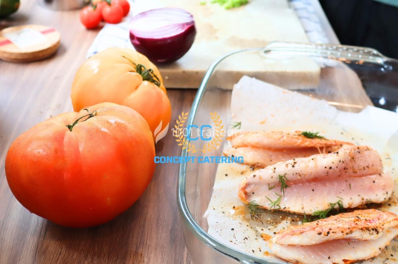 Filets de poisson marinés avec herbes dans un plat en verre, entourés de tomates et d'oignon sur un plan de travail.