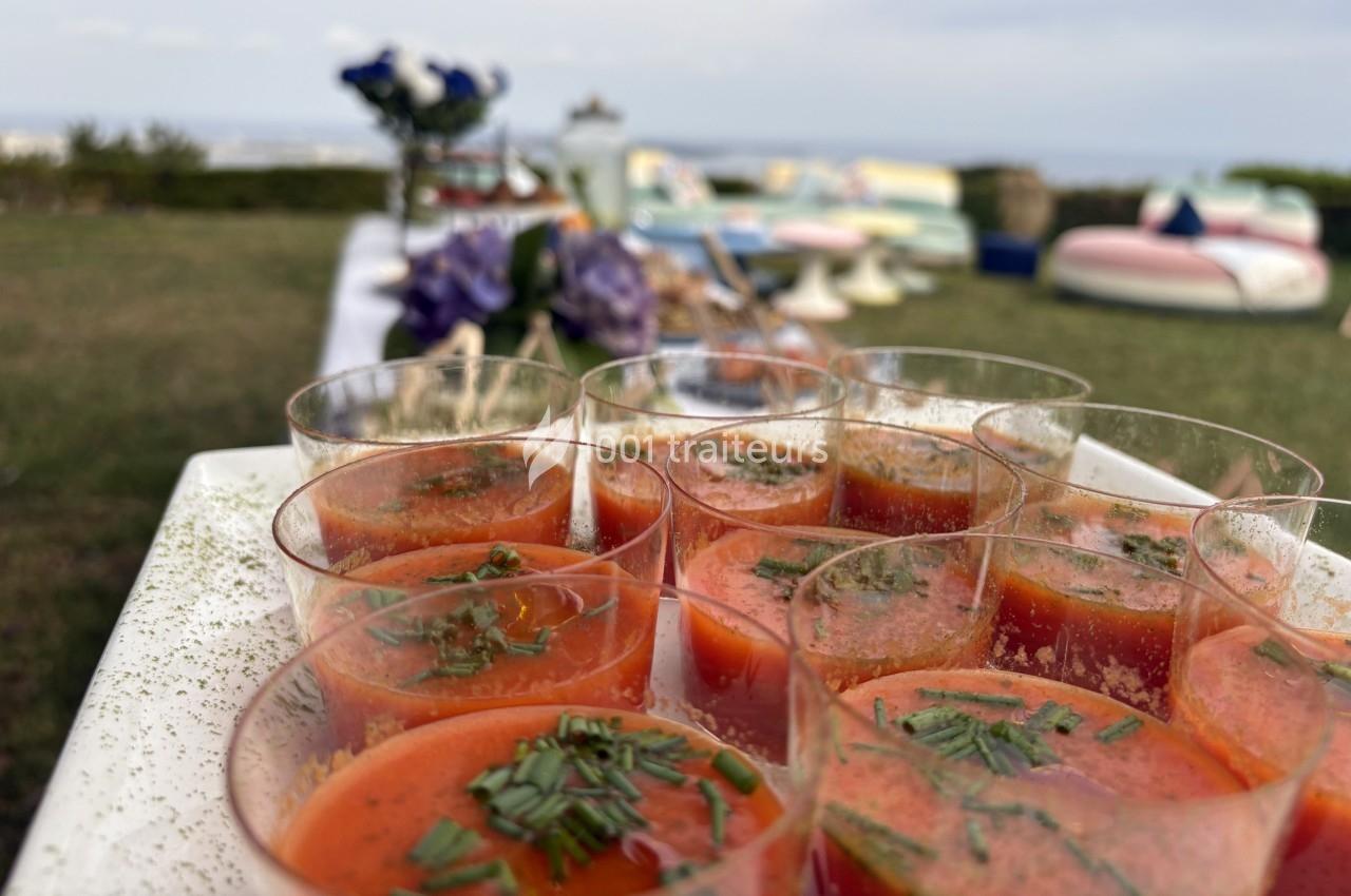 Plateau de verrines de soupe rouge garnies de ciboulette, disposé en extérieur avec un décor flou en arrière-plan.