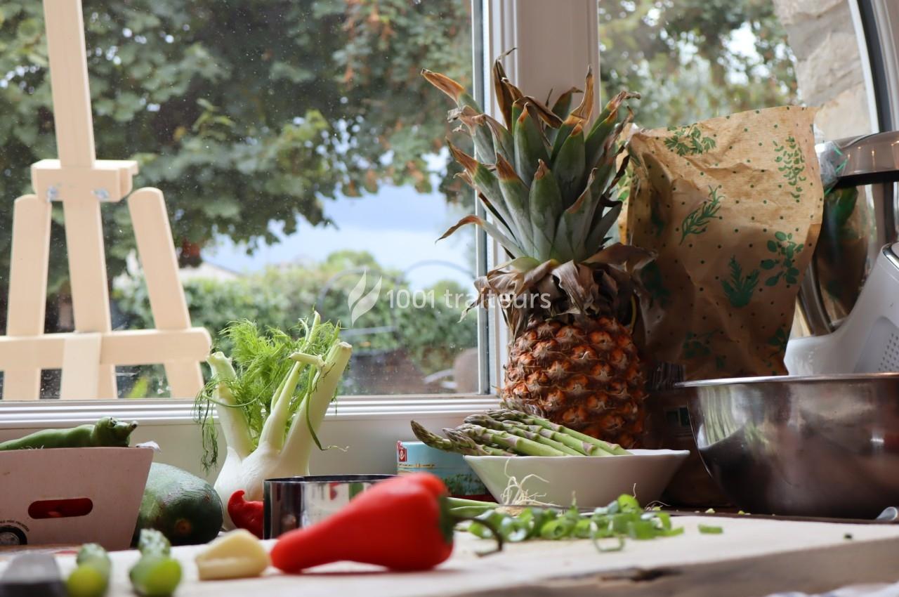 Fruits, légumes et ustensiles de cuisine disposés sur un plan de travail près d'une fenêtre donnant sur un jardin.