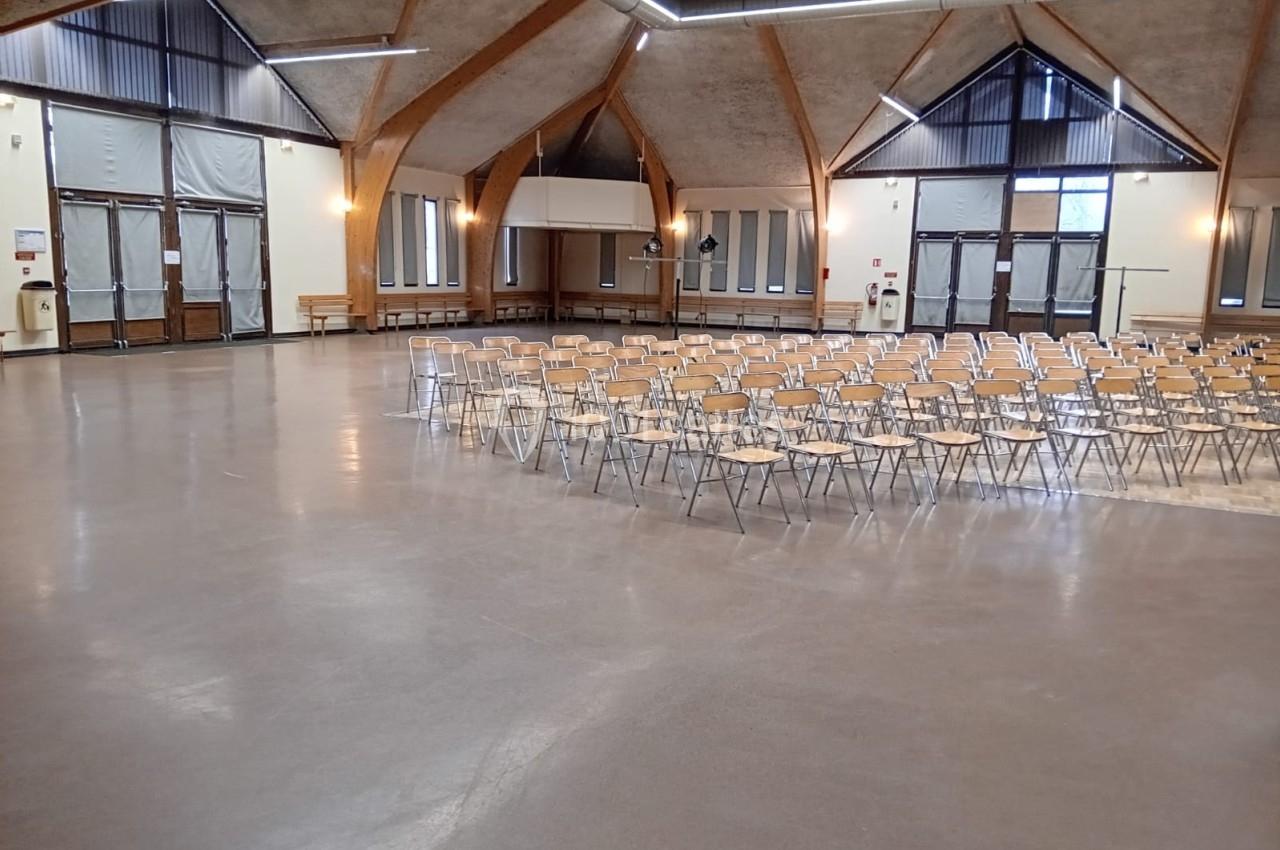 Salle spacieuse avec des rangées de chaises vides, éclairée par des fenêtres et des luminaires suspendus.