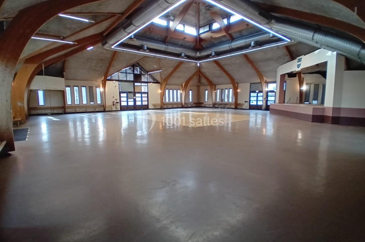Salle spacieuse avec charpente en bois apparente, grandes fenêtres et éclairage naturel provenant du plafond.