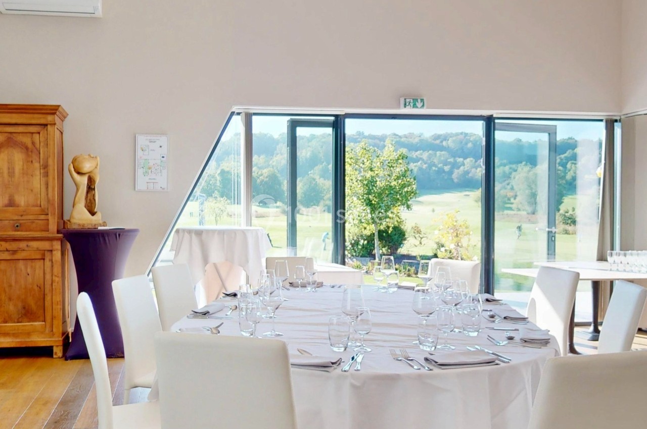Salle lumineuse avec table ronde dressée, chaises blanches, grande baie vitrée donnant sur un paysage verdoyant.