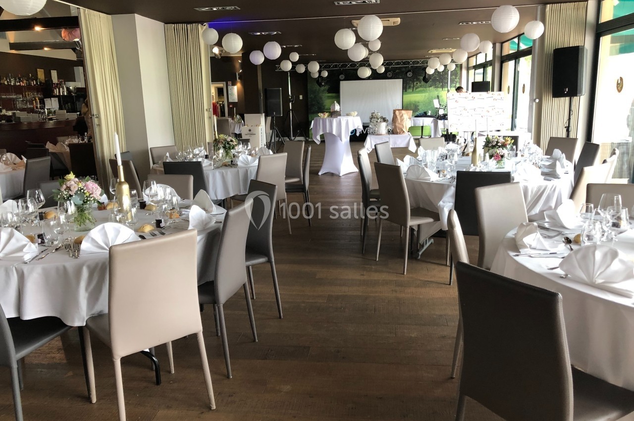 Salle de réception élégante avec tables rondes dressées, chaises modernes et décorations suspendues.