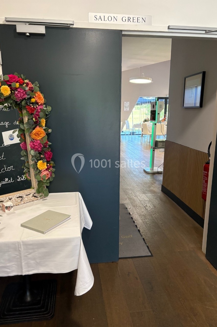 Entrée d'une salle nommée ’Salon Green’ décorée pour un mariage, avec une table et une ardoise fleurie à gauche.