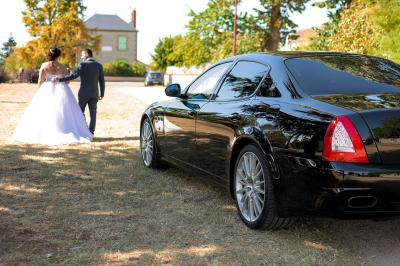 Voiture noire garée sur un chemin herbeux, avec un couple en tenue de mariage marchant au loin.