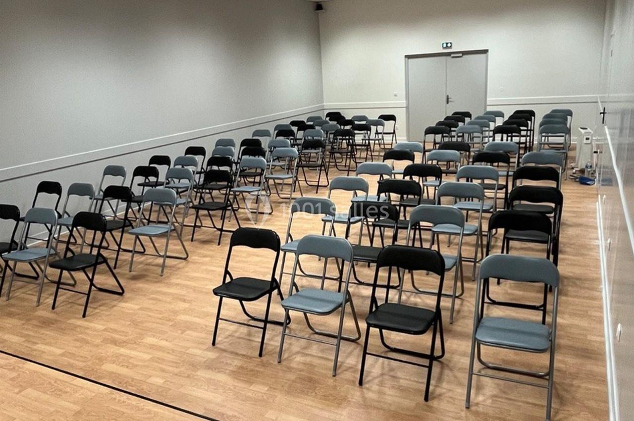Salle avec des rangées de chaises pliantes noires et grises disposées sur un sol en bois clair, éclairée par des néons.