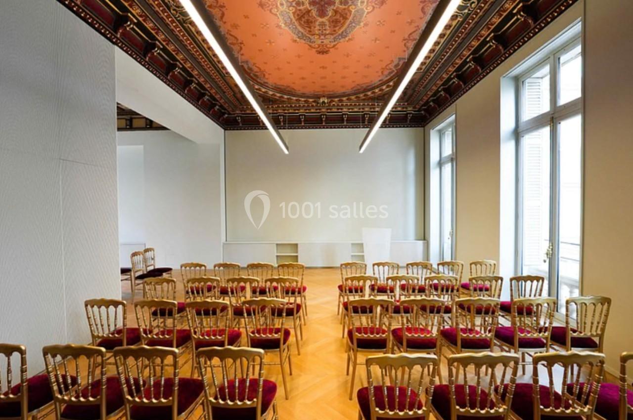 Salle de conférence lumineuse avec rangées de chaises dorées et plafond décoré de motifs rouges et dorés.