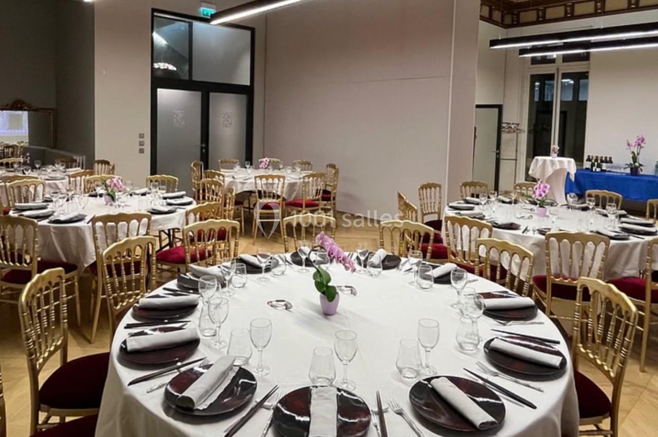 Salle de réception élégante avec tables rondes dressées, chaises dorées et décoration florale.