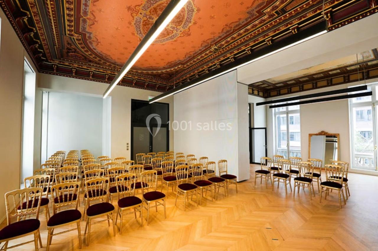 Salle lumineuse avec parquet en bois, plafond décoratif et chaises alignées pour un événement ou une réunion.
