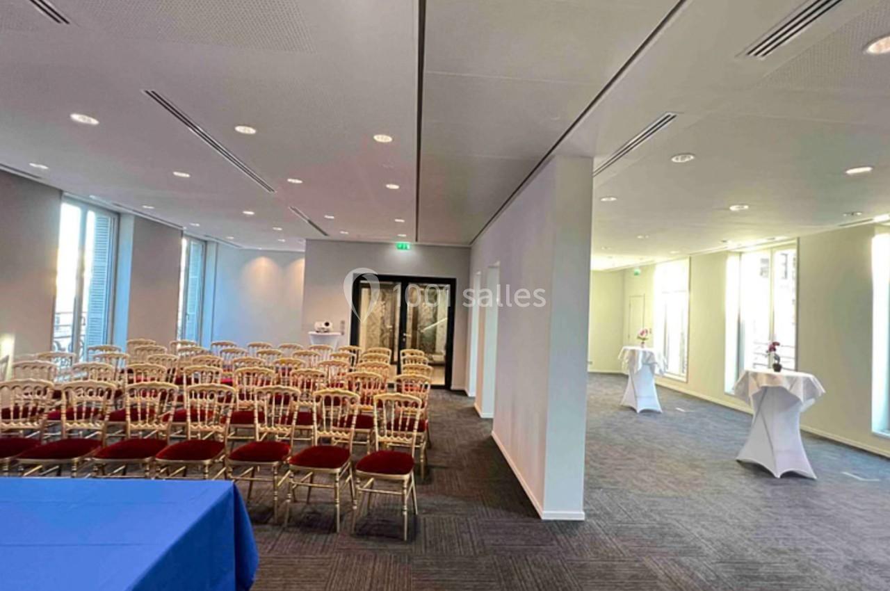 Salle de conférence lumineuse avec rangées de chaises dorées, tables hautes et grandes fenêtres laissant entrer la lumière…