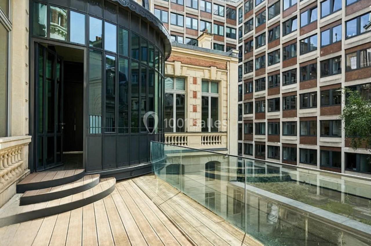 Terrasse en bois avec garde-corps en verre, donnant sur une cour intérieure entourée de bâtiments modernes et anciens.