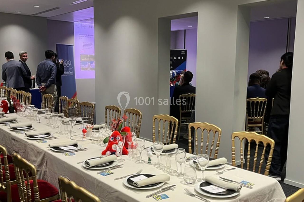 Salle de réception avec tables dressées, décorations simples et quelques personnes discutant en arrière-plan.