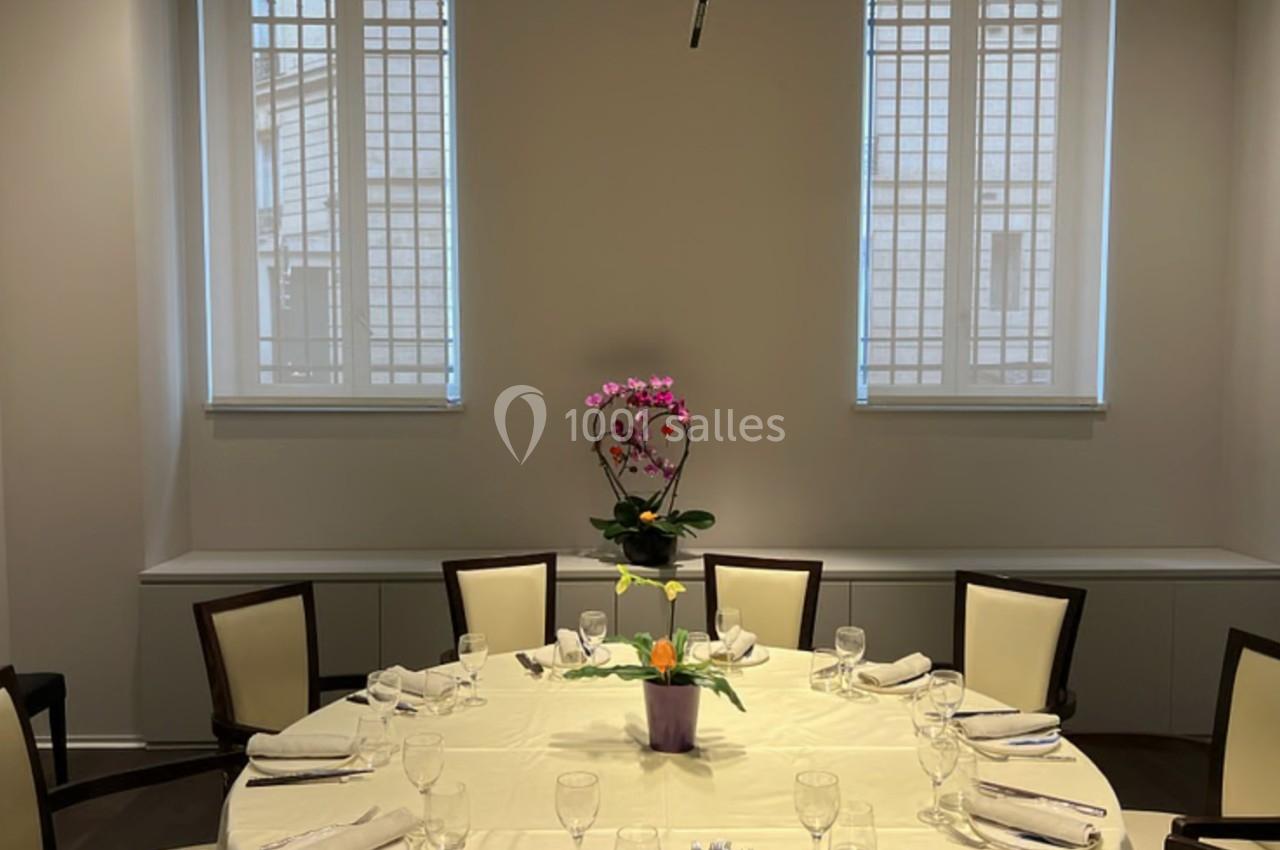 Table ronde dressée avec une nappe blanche, entourée de chaises, et ornée d'une plante au centre, dans une salle lumineuse.
