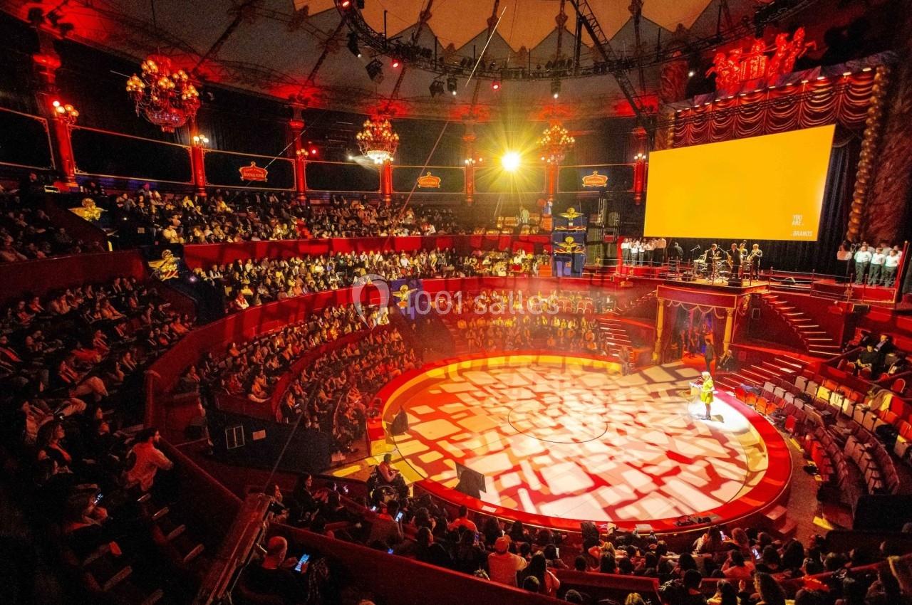 Vue intérieure d'un cirque avec une piste éclairée, des spectateurs dans les gradins et une scène en arrière-plan.