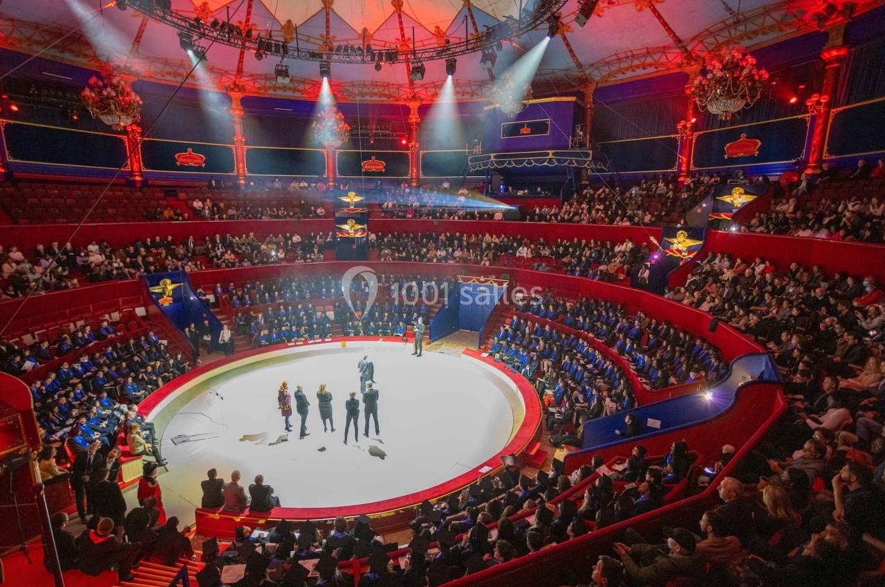Vue d'une piste de cirque entourée de gradins remplis de spectateurs, éclairée par des projecteurs colorés.