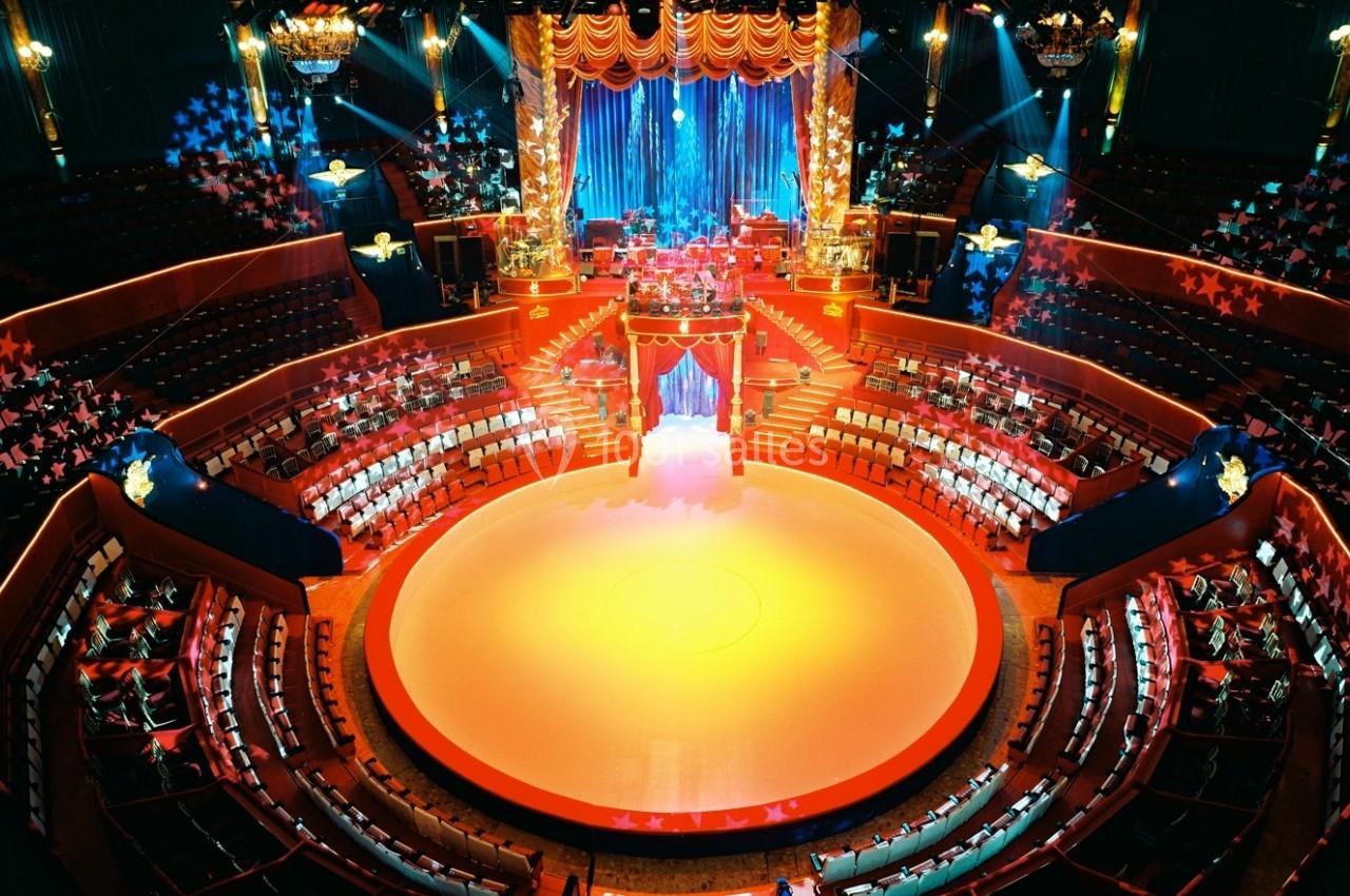 Vue intérieure d'un cirque avec piste centrale éclairée, gradins rouges et décorations lumineuses étoilées.