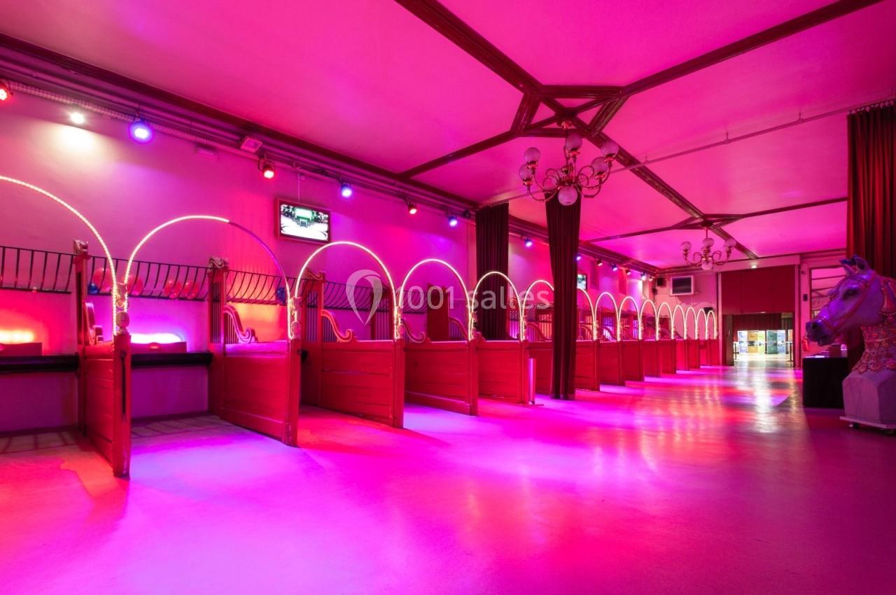 Écurie transformée en espace intérieur éclairé par des lumières roses et violettes, avec des boxes alignés.