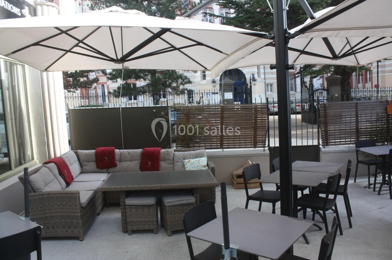 Terrasse extérieure avec mobilier en rotin, tables noires et parasols blancs, entourée de bâtiments urbains.