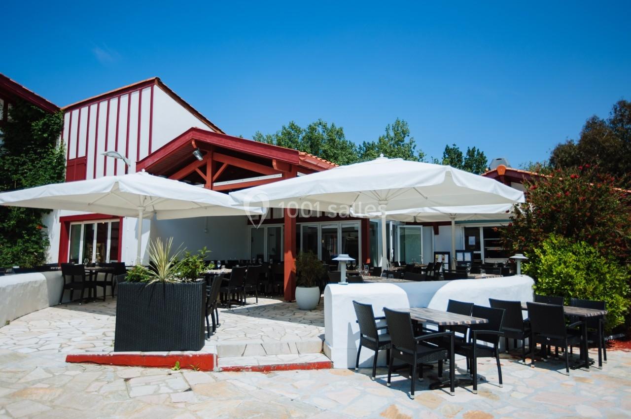 Terrasse d'un restaurant avec tables, chaises noires et grands parasols blancs, entourée de végétation sous un ciel bleu.
