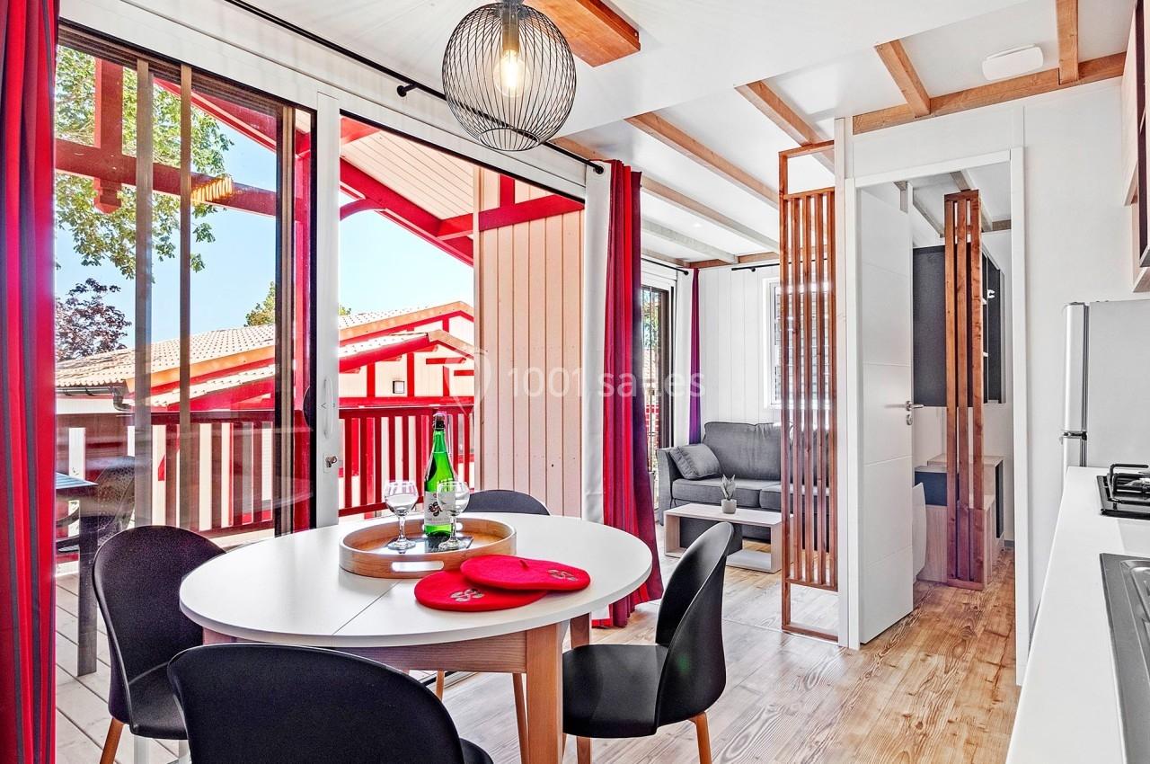 Salle à manger lumineuse avec table ronde, chaises noires, cuisine ouverte et accès à un balcon en bois.