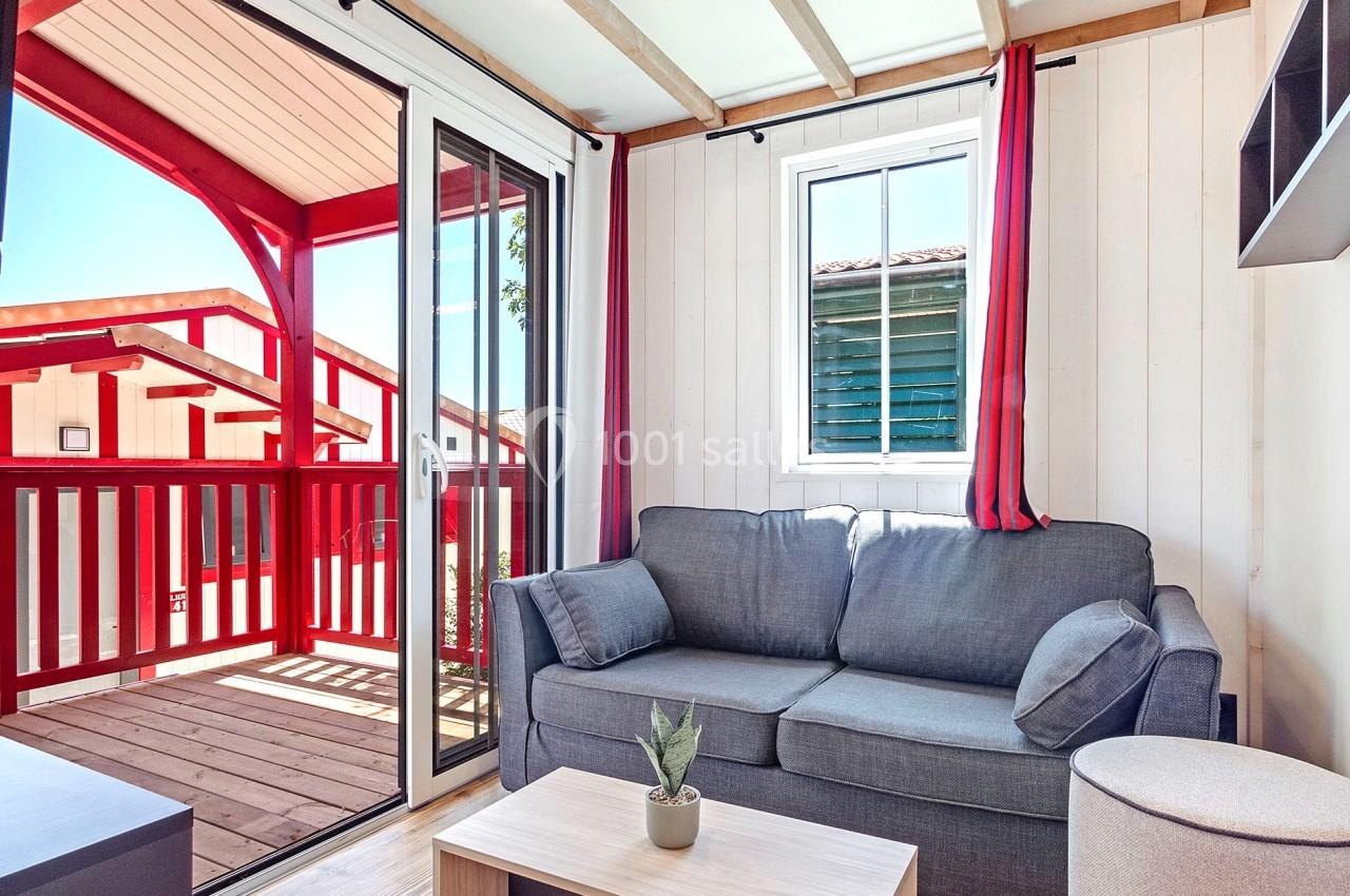 Salon lumineux avec canapé gris, table basse en bois clair, fenêtre et porte-fenêtre donnant sur une terrasse en bois rouge.