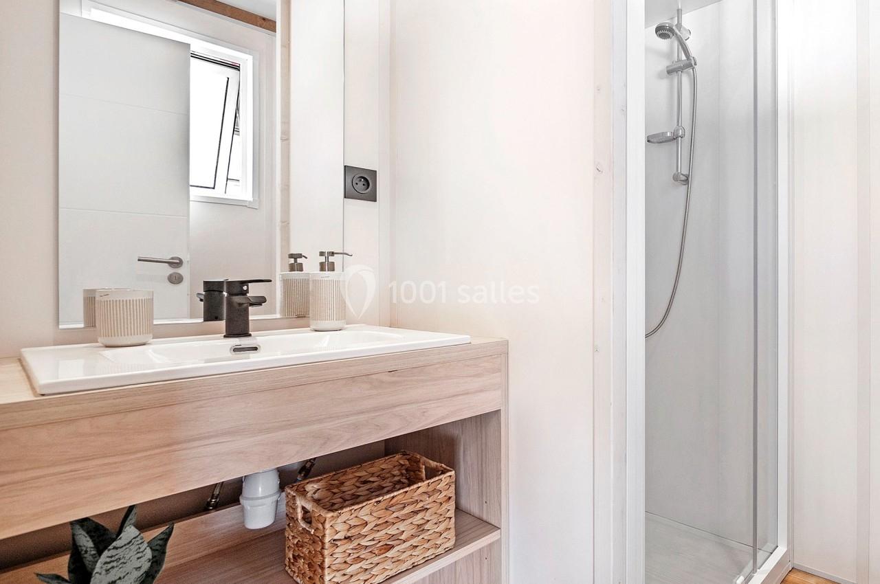 Salle de bain lumineuse avec lavabo en bois, robinet noir, panier en osier et cabine de douche vitrée.