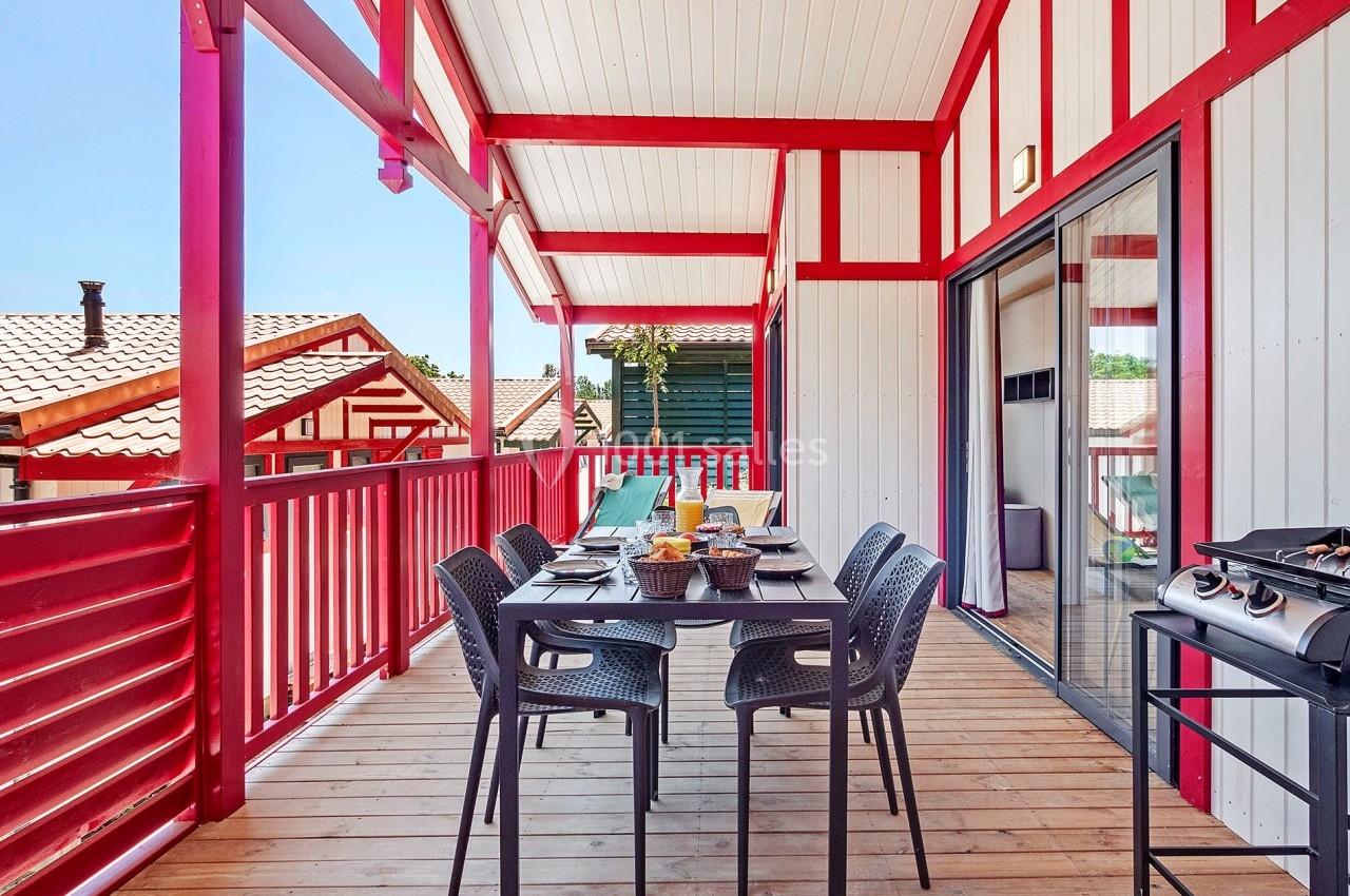 Terrasse en bois avec table dressée, chaises noires, barbecue et vue sur des toits de maisons, sous une pergola rouge.