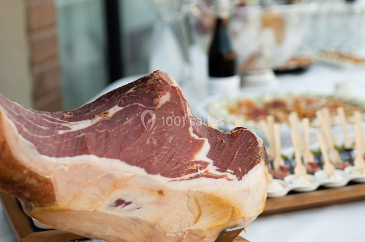 Un jambon cru sur un support en bois, avec une table garnie d'amuse-bouches et de boissons en arrière-plan.