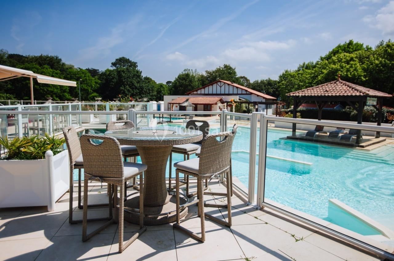 Terrasse avec table et chaises en rotin surplombant une piscine extérieure entourée de végétation et bâtiments.