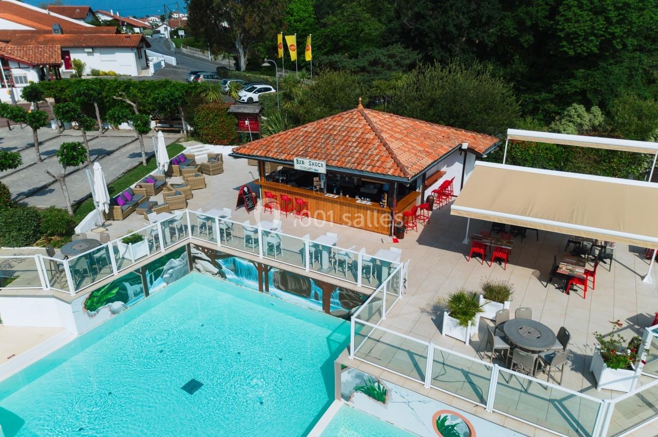 Vue aérienne d'une piscine extérieure entourée de terrasses aménagées avec un bar-restaurant en arrière-plan.