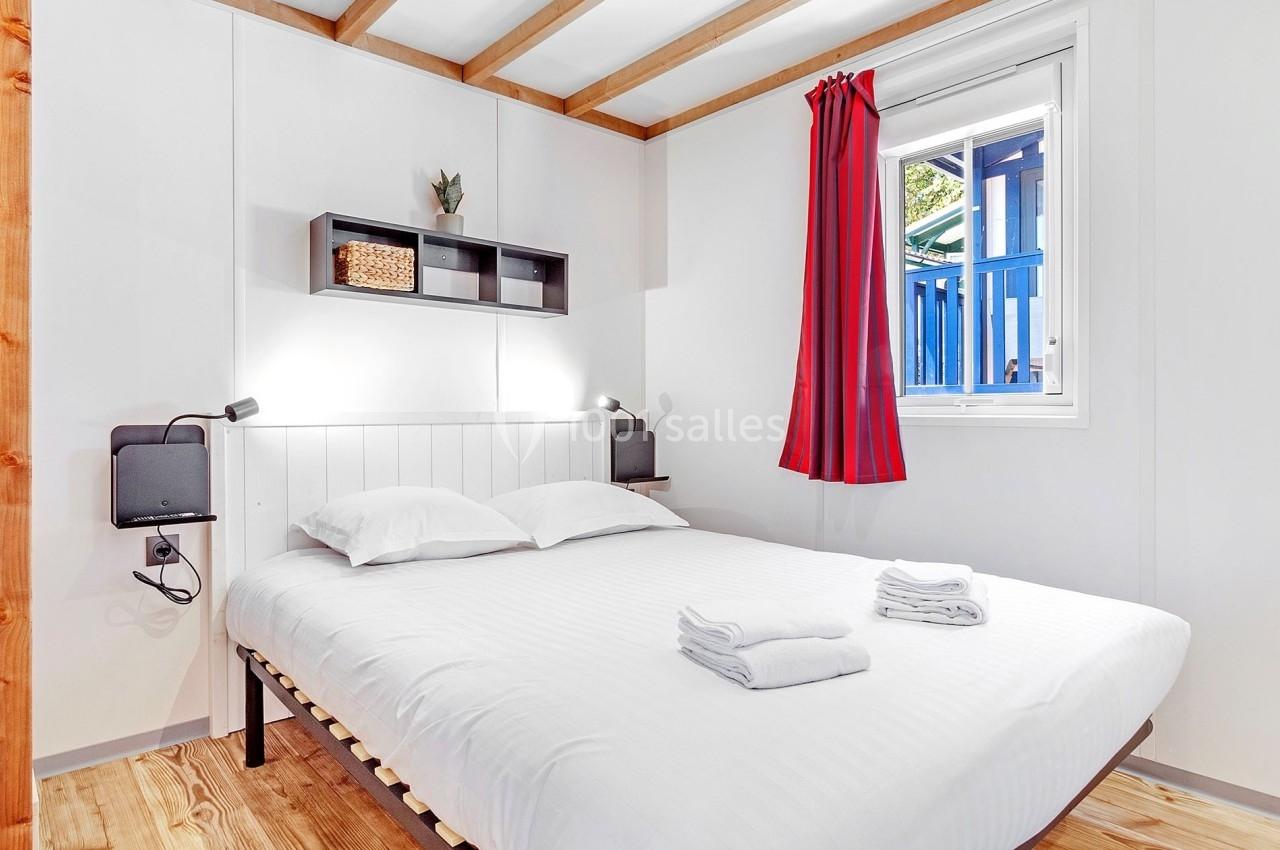Chambre lumineuse avec un lit double, draps blancs, étagère murale, fenêtre avec rideaux rouges et sol en bois.