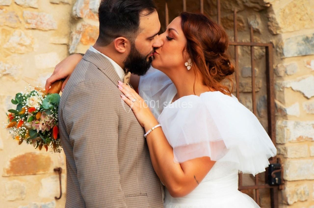 Un couple en tenue de mariage s'embrasse devant un mur en pierre, la mariée tenant un bouquet coloré.