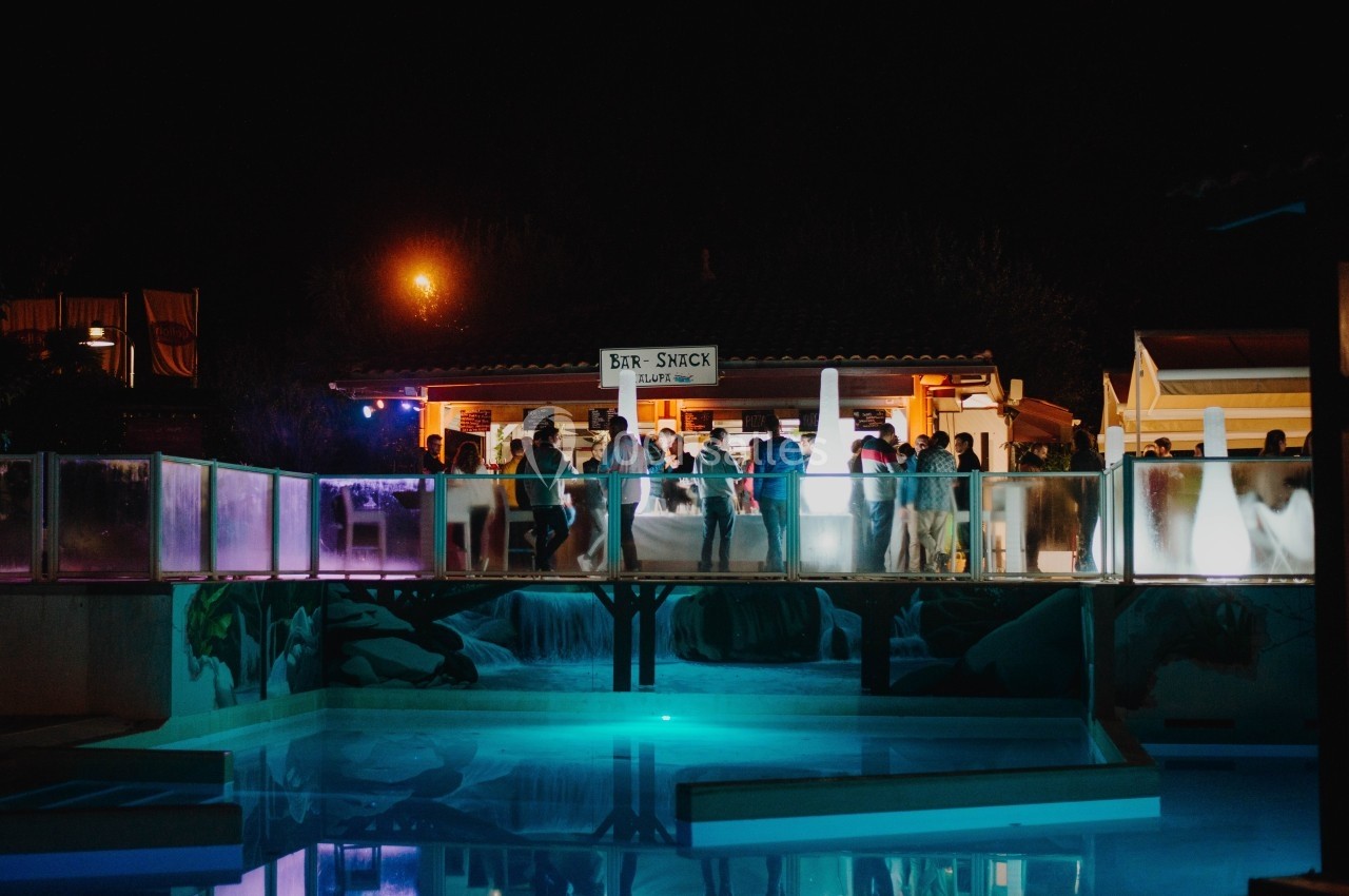 Groupe de personnes rassemblées devant un bar éclairé la nuit, avec une piscine illuminée au premier plan.