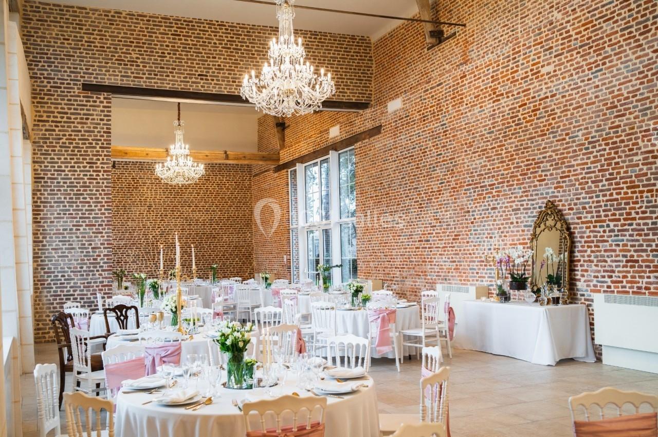 Salle de réception avec murs en briques, tables rondes dressées, chaises blanches et lustres suspendus.