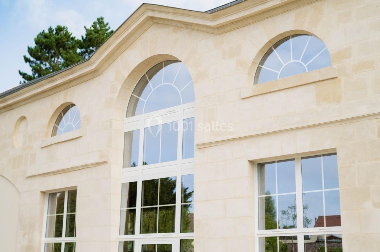 Façade d'un bâtiment en pierre claire avec de grandes fenêtres cintrées, entouré de végétation et d'un ciel bleu.