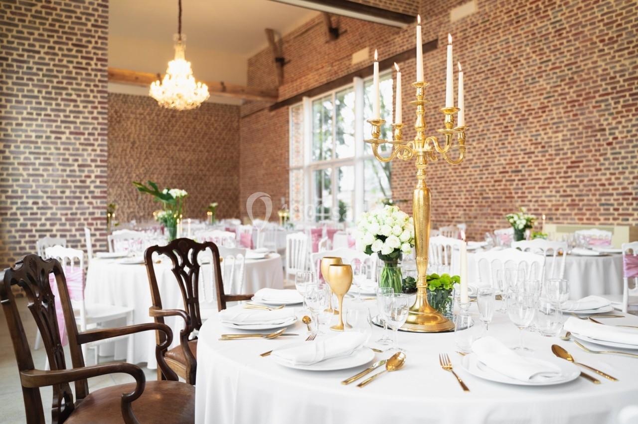 Salle de réception élégante avec tables dressées, nappes blanches, chandeliers dorés et murs en briques apparentes.