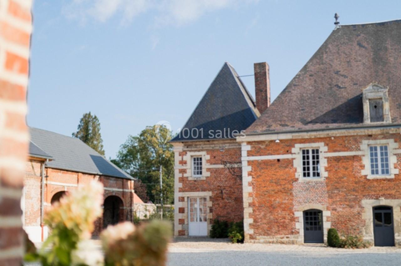 Cour pavée avec un bâtiment en briques rouges et toit en ardoise, entouré de végétation sous un ciel bleu.