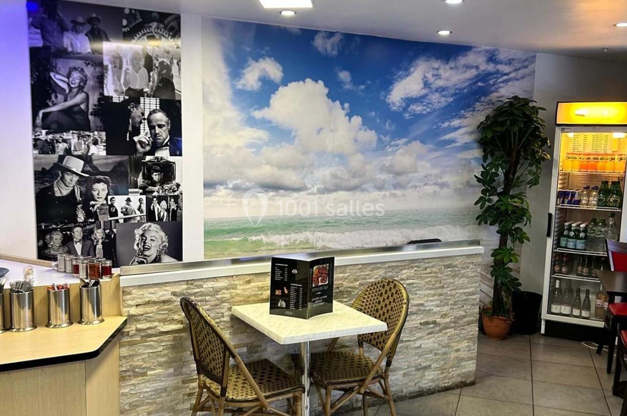 Salle de restaurant avec table, chaises en rotin, mur décoré d'une fresque de plage et photos en noir et blanc.