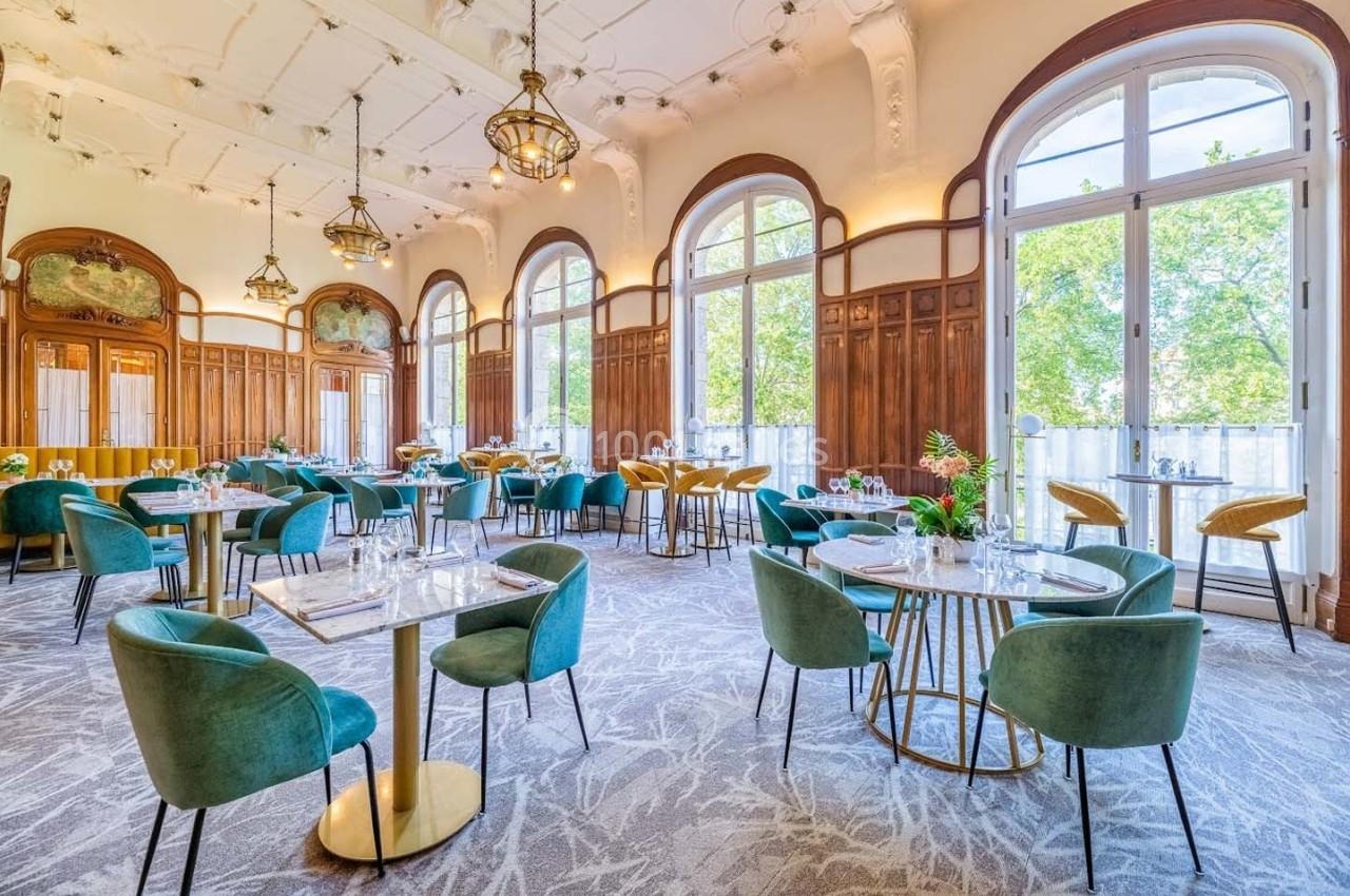 Salle lumineuse avec de grandes fenêtres, tables dressées, chaises vertes et décor élégant en bois sculpté.