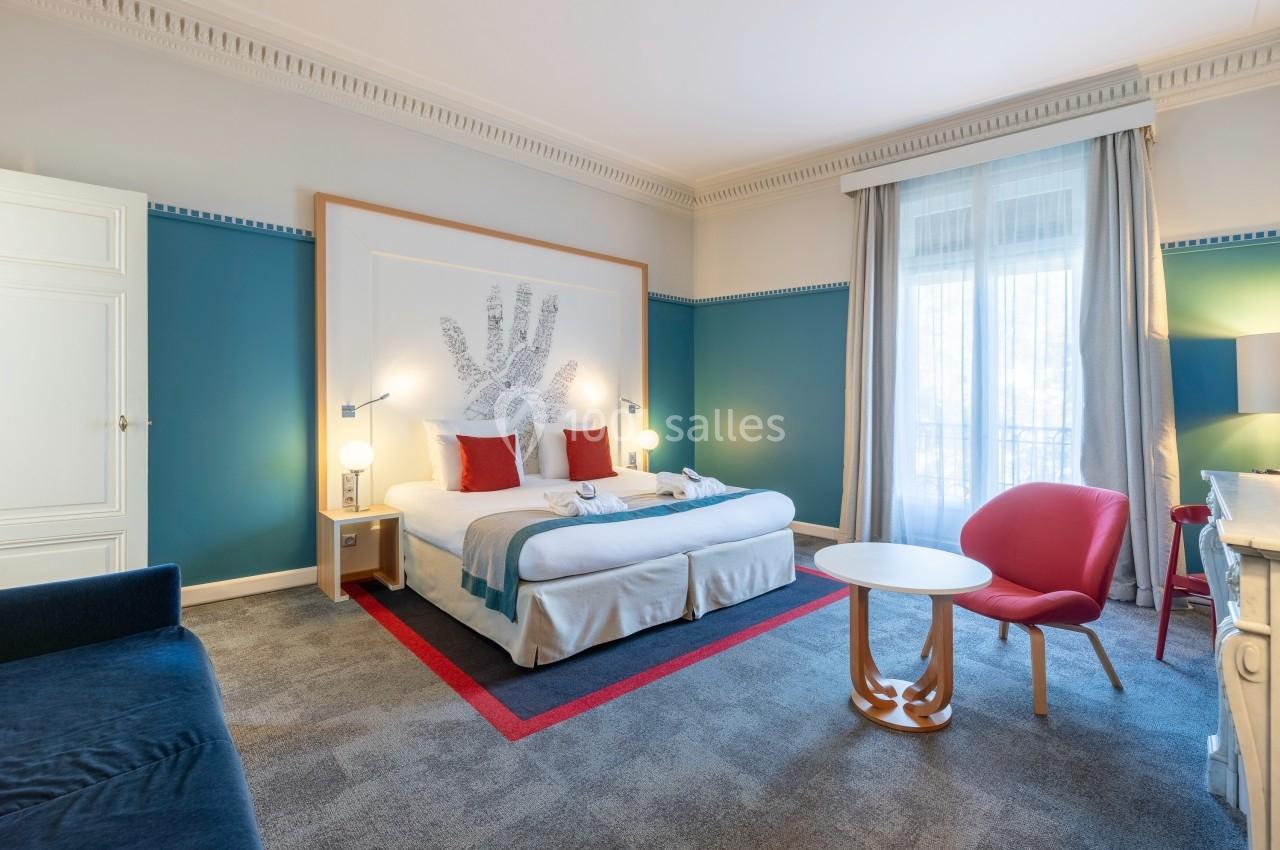 Chambre d'hôtel lumineuse avec lit double, fauteuil rouge, table ronde et grande fenêtre avec rideaux clairs.