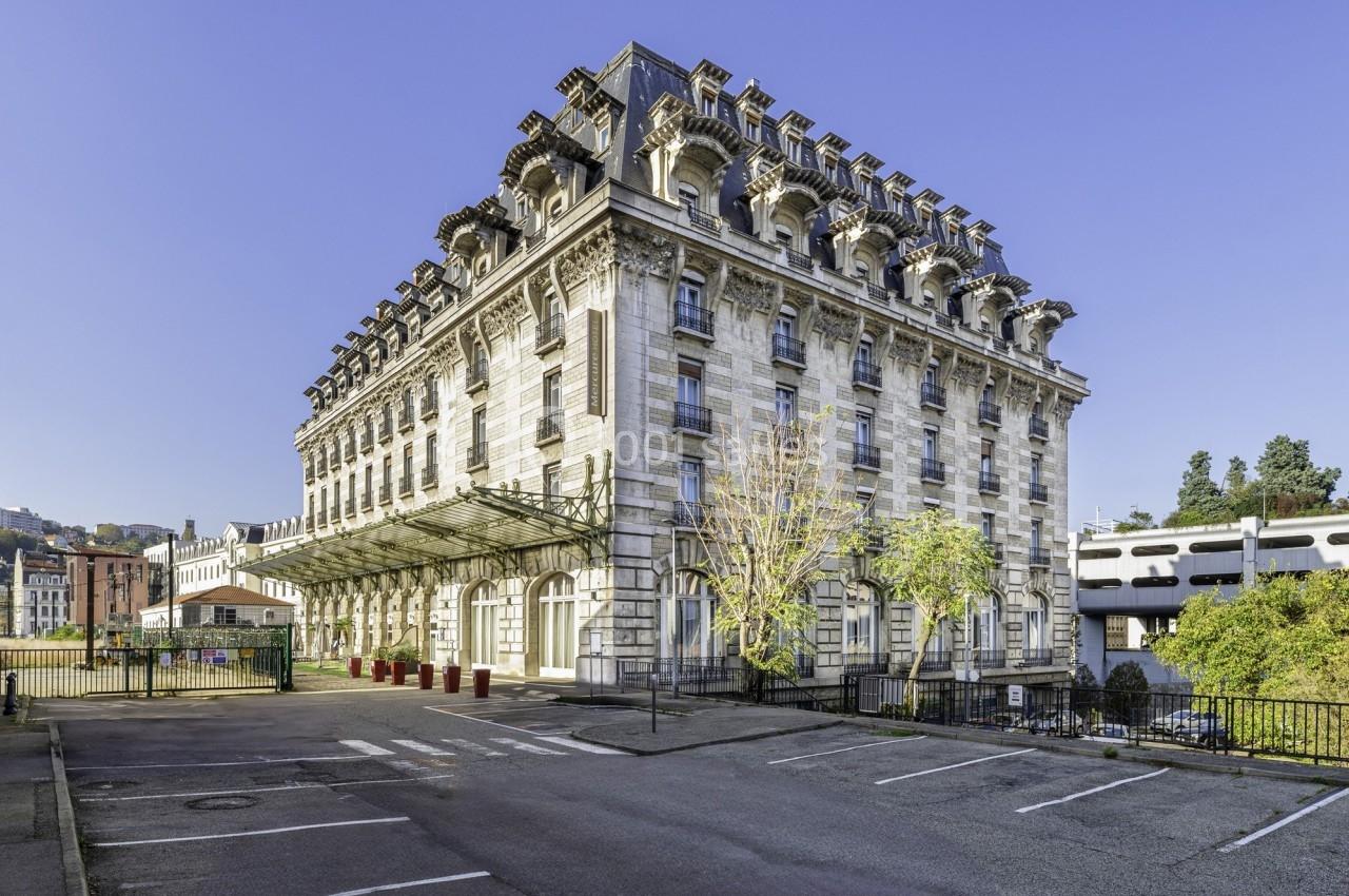 Bâtiment historique en pierre avec toit mansardé, entouré d'arbres et situé près d'un parking vide.