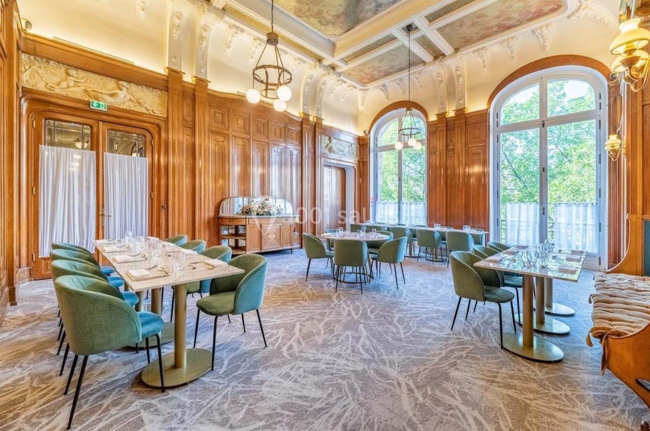 Salle lumineuse avec grandes fenêtres, murs en bois sculpté, plafond décoré et tables dressées avec chaises vertes.