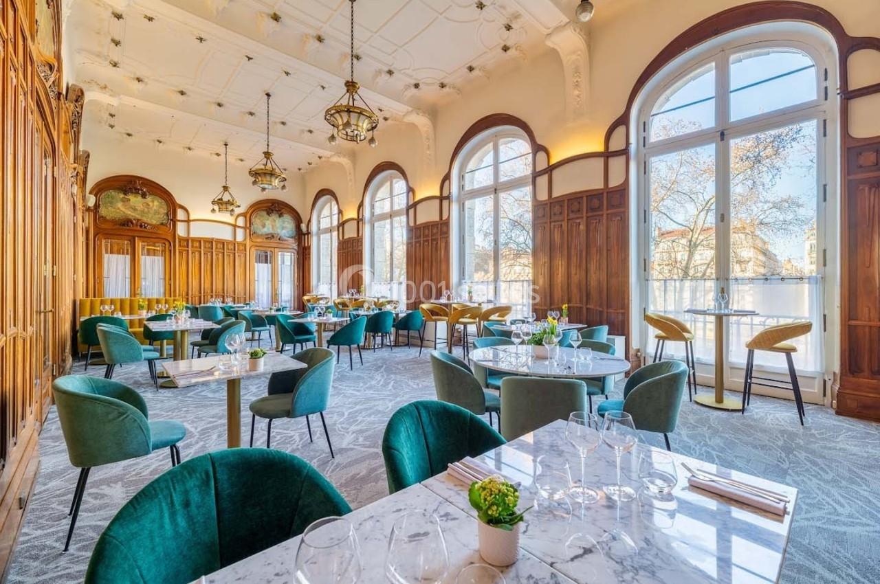 Salle de restaurant lumineuse avec grandes baies vitrées, mobilier en velours vert et tables en marbre.