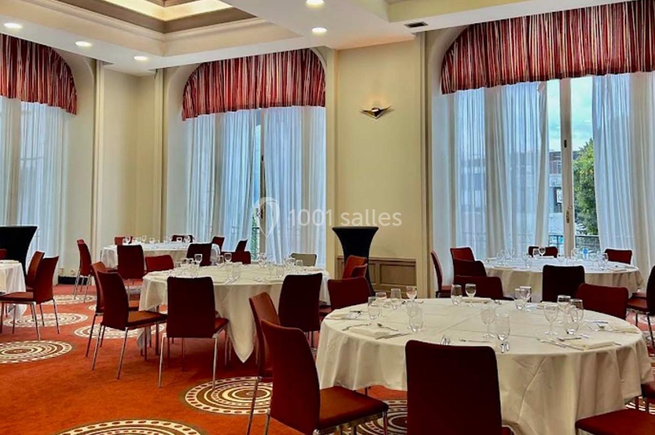 Salle de réception lumineuse avec tables rondes dressées, chaises rouges et grandes fenêtres avec rideaux.