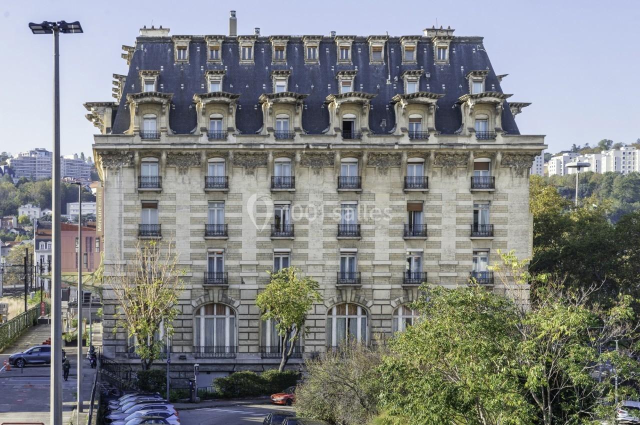 Façade d'un immeuble haussmannien avec toit mansardé, entouré d'arbres et de voitures stationnées au premier plan.