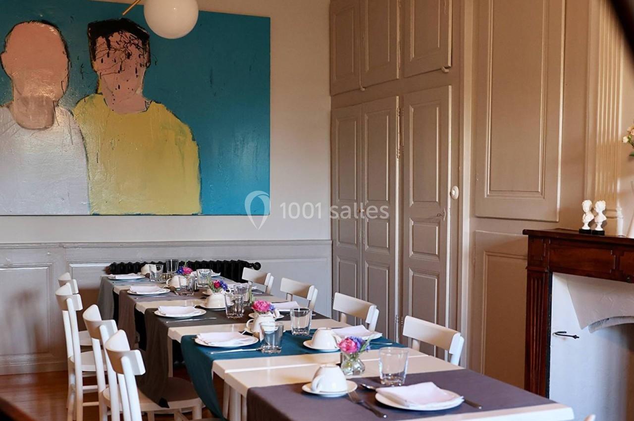 Salle à manger lumineuse avec une grande table dressée, des chaises blanches et un tableau coloré accroché au mur.