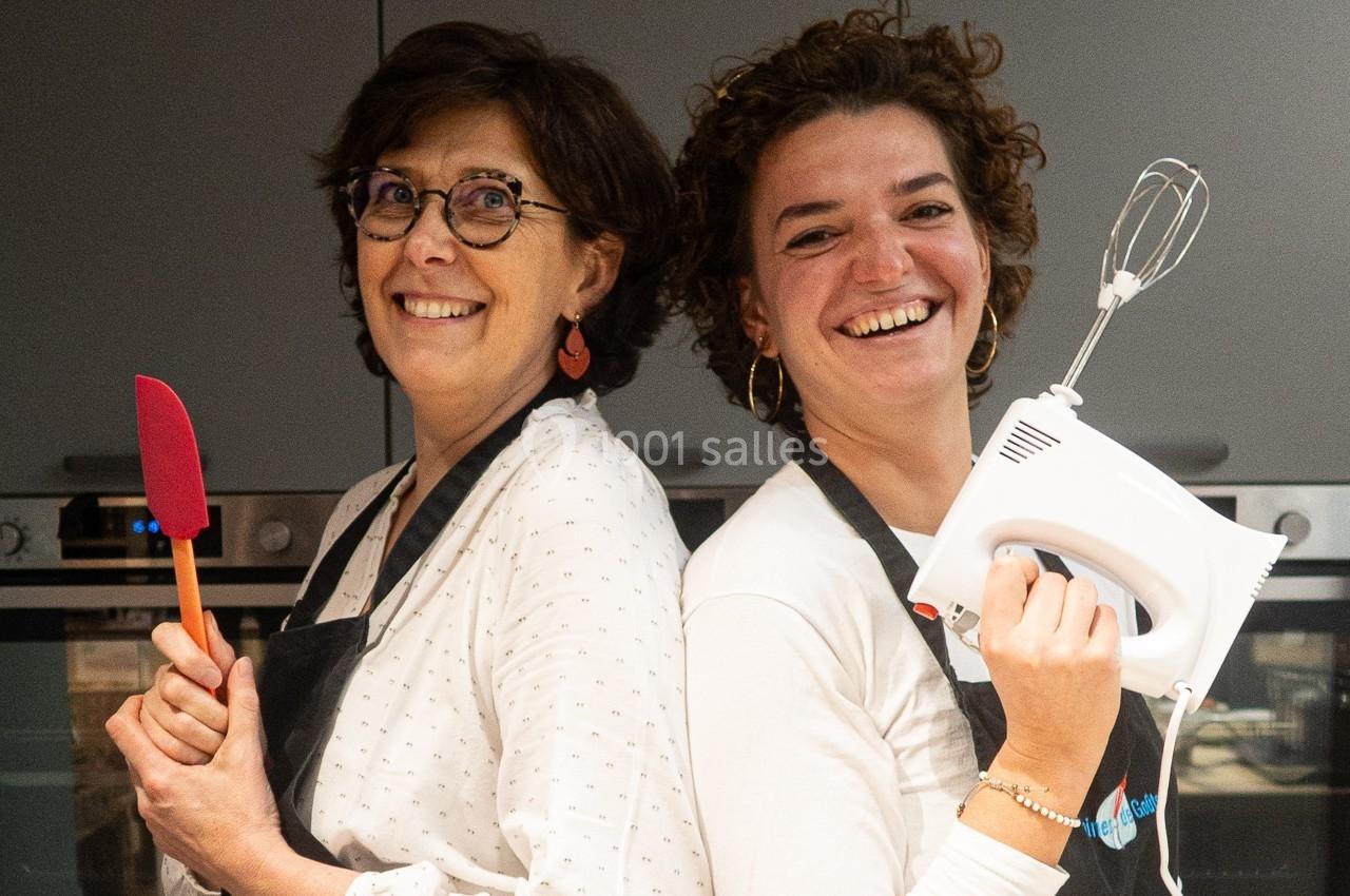 Deux femmes souriantes en tablier, l'une tenant une spatule rouge, l'autre un batteur électrique, dans une cuisine.