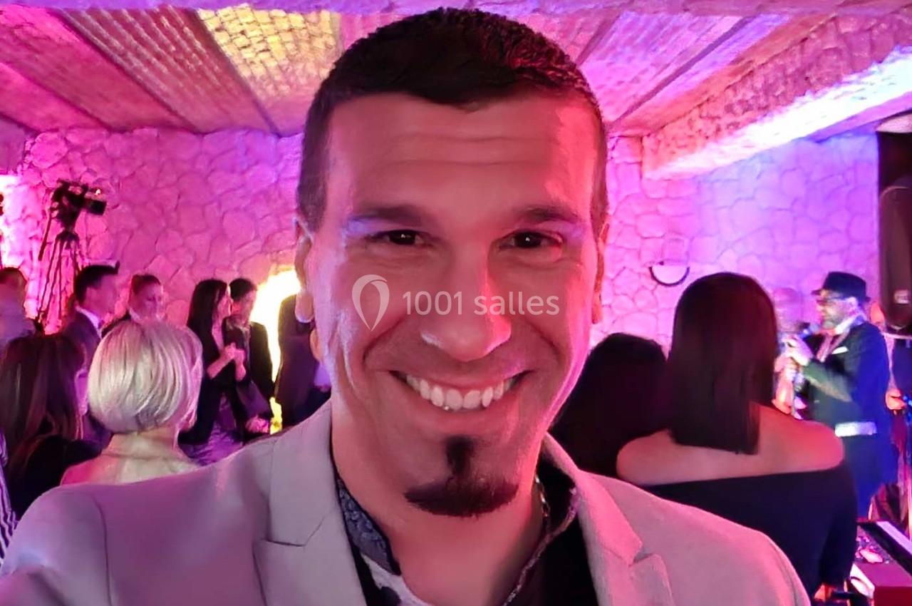 Homme souriant prenant un selfie lors d'un événement festif avec des invités et un musicien en arrière-plan.