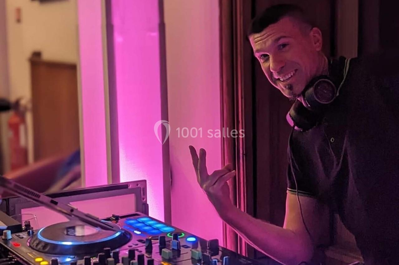 Un DJ souriant avec un casque autour du cou se tient près d'une table de mixage éclairée par une lumière rose.
